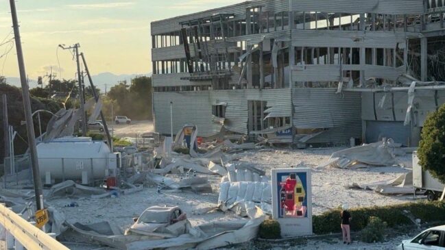 Fotografia ao nível do chão mostrando severos danos estruturais na fachada de um centro comercial de múltiplos andares (AEON Mall) após um terremoto no Japão. As placas de revestimento metálico do prédio aparecem arrancadas e contorcidas, expondo a estrutura interna. O solo à frente está coberto por detritos, pedaços de alvenaria e poeira branca, com um carro de passeio parcialmente soterrado por chapas