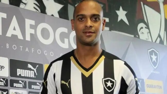 Retrato de meio-corpo do ex-jogador de futebol Tássio. Ele é um homem de pele negra, cabeça raspada e brincos ponto de luz, vestindo a camisa tradicional alvinegra (listras verticais pretas e brancas) do Botafogo, com detalhes dourados na gola V e o escudo do clube no peito esquerdo