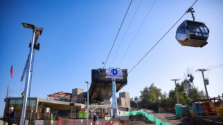 Fotografia diurna e em ângulo contra-plongé (de baixo para cima) mostrando o canteiro de obras de uma estação do Teleférico Bicentenario em Santiago. No canto superior direito, uma cabine envidraçada e moderna do teleférico está suspensa nos cabos de aço contra um céu azul sem nuvens. No centro, ergue-se uma grande estrutura metálica de sustentação com um banner branco exibindo a letra "D" azul e a marca "Doppelmayr".