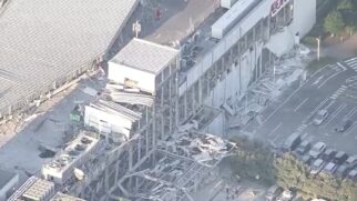 Fotografia aérea diurna mostrando os danos estruturais causados por um terremoto em um grande complexo comercial no Japão. No topo da fachada branca de um edifício, lê-se o letreiro "AEON MALL". Abaixo e ao lado, partes do teto, passarelas e paredes desabaram, acumulando escombros e retalhos metálicos na área externa.