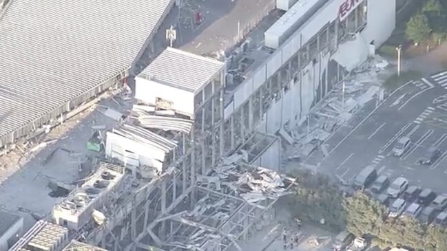 Fotografia aérea diurna mostrando os danos estruturais causados por um terremoto em um grande complexo comercial no Japão. No topo da fachada branca de um edifício, lê-se o letreiro "AEON MALL". Abaixo e ao lado, partes do teto, passarelas e paredes desabaram, acumulando escombros e retalhos metálicos na área externa.