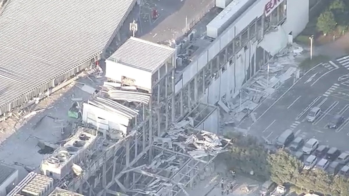Fotografia aérea diurna mostrando os danos estruturais causados por um terremoto em um grande complexo comercial no Japão. No topo da fachada branca de um edifício, lê-se o letreiro "AEON MALL". Abaixo e ao lado, partes do teto, passarelas e paredes desabaram, acumulando escombros e retalhos metálicos na área externa.