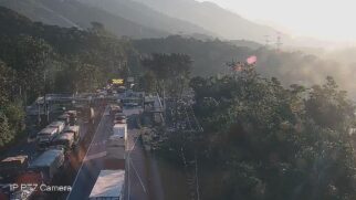 Fotografia de câmera de monitoramento rodoviário em ângulo elevado mostrando intenso congestionamento de caminhões e carretas na Rodovia Anchieta. Os veículos de carga ocupam as faixas da esquerda e do centro, formando uma longa fila antes de um pórtico com painel eletrônico de mensagem e cabines.