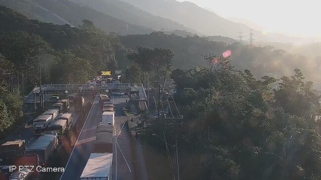 Fotografia de câmera de monitoramento rodoviário em ângulo elevado mostrando intenso congestionamento de caminhões e carretas na Rodovia Anchieta. Os veículos de carga ocupam as faixas da esquerda e do centro, formando uma longa fila antes de um pórtico com painel eletrônico de mensagem e cabines.