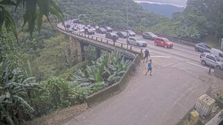 Captura de câmera de monitoramento em ângulo elevado mostrando um intenso congestionamento em um viaduto da Rodovia dos Imigrantes. Uma longa fila de carros de passeio ocupa todas as faixas da pista, que faz uma curva em meio à densa vegetação da Serra do Mar. Ao fundo, a fila se estende até a saída de um túnel
