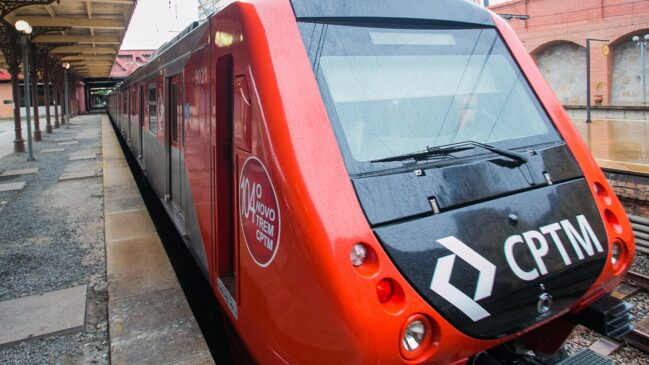 Fotografia em ângulo de três quartos de um trem moderno de cor vermelha e preta da CPTM parado ao lado da plataforma de uma estação ferroviária histórica. Na frente estilizada em tom preto, destaca-se o símbolo e a inscrição da CPTM em branco.