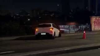 Fotografia noturna em plano médio mostrando um carro de passeio hatch branco acidentado sobre a pista de um viaduto. O veículo está parado na transversal, próximo à grade de proteção metálica da via, com o pisca-alerta e lanternas traseiras acesas. À direita, um cone de trânsito laranja e branco sinaliza o desvio para os demais motoristas.