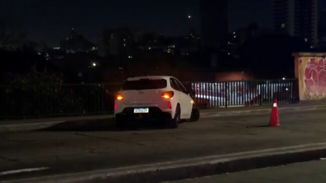 Fotografia noturna em plano médio mostrando um carro de passeio hatch branco acidentado sobre a pista de um viaduto. O veículo está parado na transversal, próximo à grade de proteção metálica da via, com o pisca-alerta e lanternas traseiras acesas. À direita, um cone de trânsito laranja e branco sinaliza o desvio para os demais motoristas.