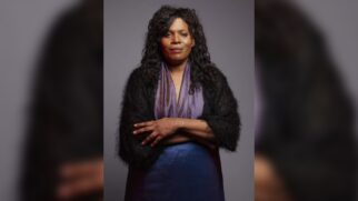 Retrato de estúdio da atriz Clodd Dias, uma mulher negra de cabelos escuros, cacheados e volumosos na altura dos ombros. Ela está com os braços cruzados na altura do peito e olha de forma serena para a câmera. Veste um vestido em tom azul-arroxeado acetinado e um xale felpudo escuro cobrindo os ombros. O fundo da imagem é neutro, em tom cinza-escuro iluminado ao centro
