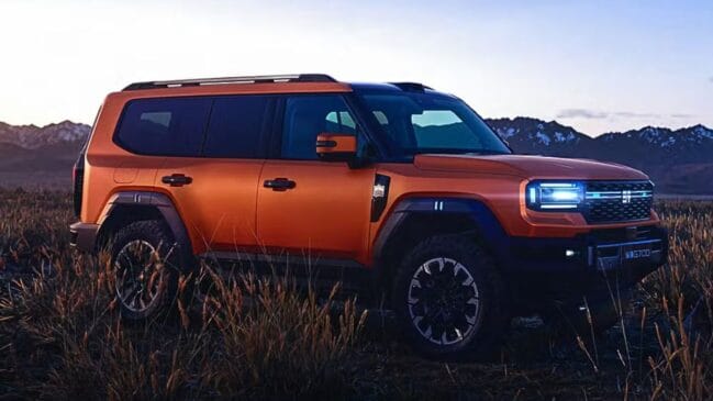 Fotografia em ângulo três-quartos dianteiro de um SUV off-road de linhas quadradas na cor laranja metálico, estacionado em um campo de vegetação baixa com montanhas ao fundo sob iluminação de crepúsculo. O veículo apresenta teto e colunas na cor preta, racks de teto, caixas de roda salientes em acabamento plástico escuro, pneus para uso em todo terreno e grade frontal com conjunto óptico em LED horizontal aceso. A placa dianteira exibe a identificação "G700"