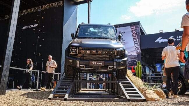 Fotografia em ângulo contra-plongée (de baixo para cima) de um SUV off-road preto, modelo Jetour T2 4x4, posicionado sobre uma rampa metálica de demonstração em um evento ao ar livre. O veículo apresenta design robusto e quadrado, com a marca "JETOUR" em destaque na grade frontal e a placa decorativa "T2 4x4". Ao fundo, do lado esquerdo, observa-se uma parede de escalada com a marca Jetour e pessoas caminhando sobre o cascalho; do lado direito, há painéis publicitários com menções à "GWM TANK 300" e "TERRAFORCE" sob céu ensolarado
