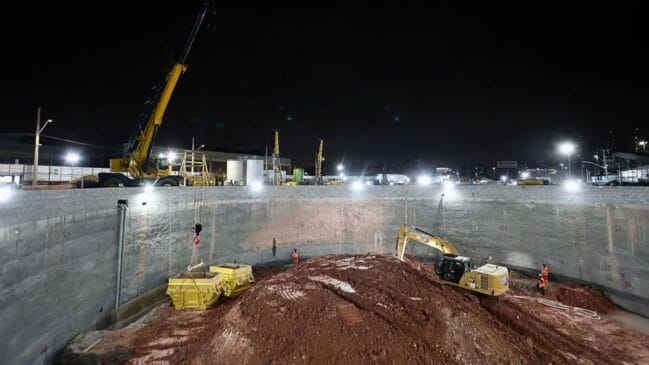 Fotografia em plano geral e iluminação noturna por refletores mostrando o interior de um grande poço circular de escavação com paredes de contenção em concreto. No fundo da escavação, uma escavadeira hidráulica amarela atua sobre um monte de terra avermelhada perto de operários em trajes de proteção e coletes alaranjados. Na borda superior do poço, um guindaste amarelo eleva uma caçamba metálica de entulho carregada de terra. Ao fundo, sob o céu escuro da noite, observam-se outras máquinas e equipamentos do canteiro de obras