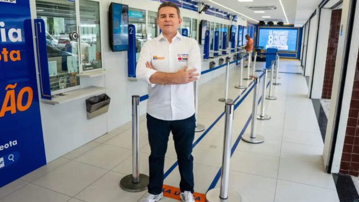 Fotografia em plano inteiro capturada em ambiente interno e bem iluminado, mostrando um homem de cabelos castanhos, vestindo camisa social branca com o logotipo "Rei do Bolão", calça jeans escura e tênis claros, de pé e com os braços cruzados no corredor central de uma casa lotérica. Ao fundo, veem-se os guichês de atendimento com vidros numerados, pedestais organizadores de fila com fitas azuis e um painel digital azul com a frase "Já pagamos 8 prêmios principais".