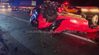 Fotografia noturna em plano médio mostrando um carro de passeio vermelho totalmente capotado de rodas para cima no asfalto de uma rodovia. O veículo apresenta amassados na lataria, porta aberta e estilhaços espalhados pela pista junto ao acostamento. Ao fundo, veem-se uma viatura de resgate vermelha e amarela com luzes de emergência ligadas, um caminhão guincho da concessionária e marcas d'água transparentes com a sigla "ARTESP"