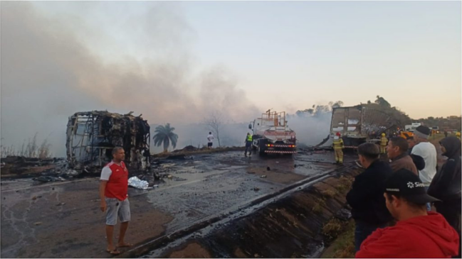 otografia em plano geral e iluminação diurna mostrando o cenário de um grave acidente com incêndio na rodovia BR-040. À esquerda, observa-se a carcaça completamente carbonizada e destruída de um ônibus, com fumaça subindo ao fundo. No centro da pista coberta por fuligem e água, um caminhão-pipa branco atua no rescaldo. À direita, os restos queimados de um caminhão ocupam a margem da via perto de bombeiros com capacetes amarelos. Em primeiro plano, moradores e motoristas observam a cena a partir da canaleta lateral da pista