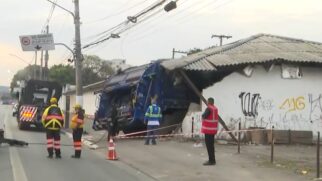 Fotografia no nível da rua ao amanhecer mostrando um caminhão azul de coleta de lixo colidido contra o muro e o telhado de um imóvel térreo de parede branca. O impacto causou o desabamento parcial do telhado de telhas onduladas e danos estruturais na parede