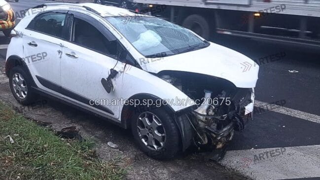 Fotografia em plano três-quartos dianteiro de um veículo hatch de cor branca acidentado e parado sobre a sarjeta do acostamento em uma rodovia. O automóvel apresenta severos danos na extremidade dianteira direita, com o para-choque destruído, estrutura do motor exposta, teto e para-lama amassados, retrovisor lateral pendurado e para-brisa trincado. O airbag frontal do passageiro está deflagrado e visível pela janela. Na parte inferior, há uma estampa de data e hora registrada como "26 de ago. de 2026 07:23:42" e marcas d'água com o logotipo da Artesp sobrepostas na imagem. Ao fundo, um caminhão passa pela faixa de rolamento