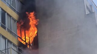 Fotografia aproximada em ângulo inferior mostrando chamas intensas irrompendo da janela de um apartamento em prédio residencial. Labaredas altas nas cores laranja e amarelo saem pela caixilharia da janela, enquanto uma densa fumaça preta sobe ao longo da fachada do edifício. À direita, vê-se a parede cega de concreto cinza do prédio vizinho envolta por uma névoa de fumaça