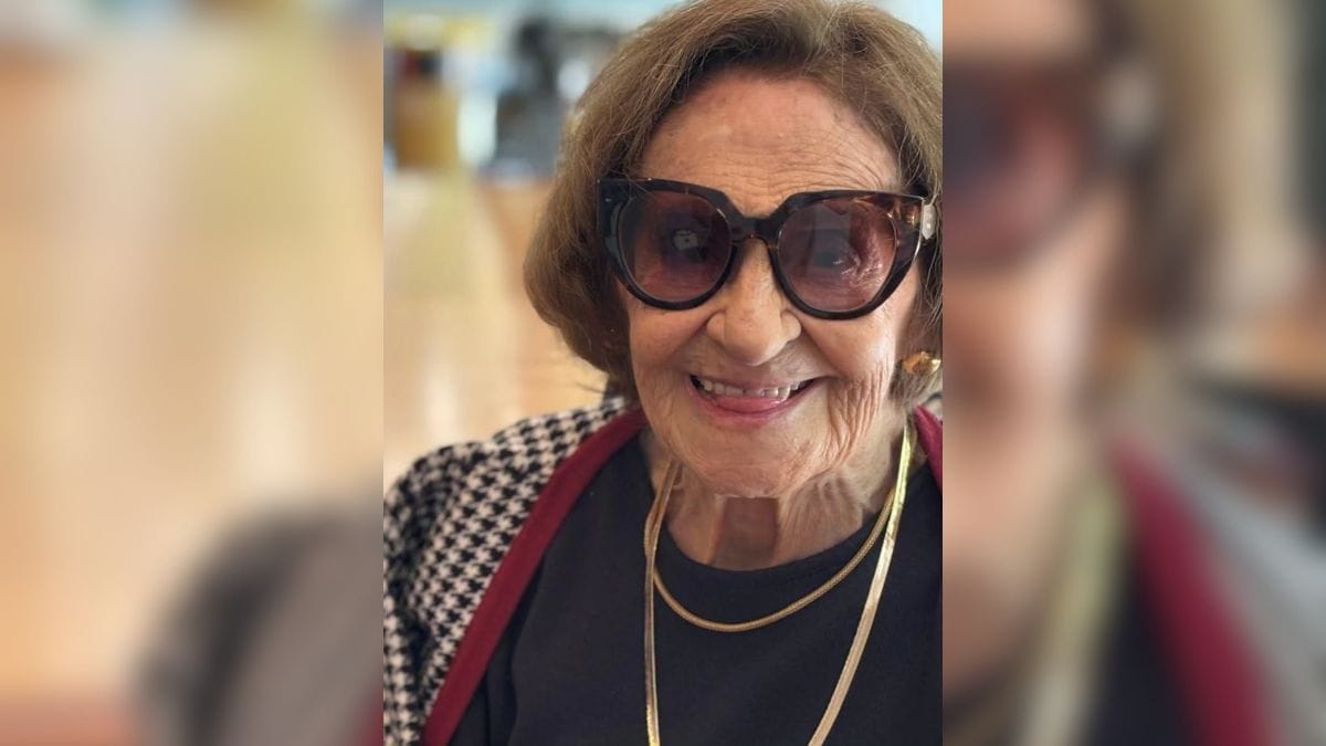 Fotografia em primeiro plano da atriz veterana Laura Cardoso sorrindo. Ela veste blusa preta, casaco com padronagem xadrez em preto e branco com bordas vermelhas, colar de fita dourado e óculos de sol de armação grande estilo gatinho com estampa de tartaruga. Ao fundo, observa-se um ambiente interno iluminado e desfocado
