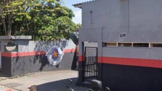 Fotografia de rua em plano médio mostrando a fachada e o portão de acesso de uma unidade da Polícia Militar do Estado de São Paulo (PMESP). O prédio é pintado nas cores cinza e preta com uma faixa horizontal vermelha central e traz o número 270 acima da entrada de grade preta. No muro à esquerda, destaca-se o brasão oficial da PMESP pintado em grandes dimensões. Ao fundo, observam-se árvores sob iluminação diurna