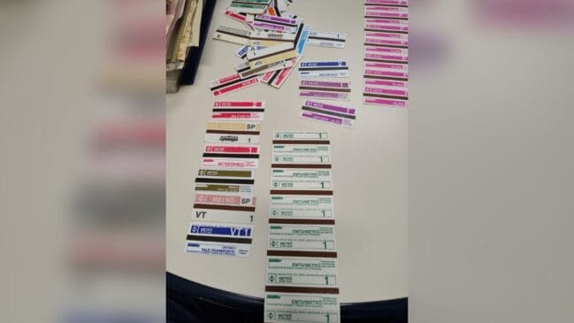 Fotografia vista de cima de uma coleção de antigos bilhetes magnéticos de papel do Metrô de São Paulo organizados sobre uma mesa branca. Os bilhetes retangulares apresentam a clássica tarja magnética escura e diferentes layouts coloridos com inscrições como "Unitário", "Idoso", "Vale-Transporte", "EMTU/Metrô", "Ida e Volta" e "Unitário Lilás". Os tíquetes estão dispostos em colunas alinhadas no centro e em uma pilha desordenada no topo da imagem sob luz ambiente de escritório