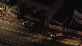 Fotografia aérea noturna mostrando a cena de uma ocorrência policial na Zona Norte de São Paulo. Duas viaturas da Polícia Militar (PM-SP) estão estacionadas na calçada, com luzes de emergência ligadas, em frente a edifícios comerciais escuros