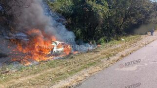 Fotografia diurna no acostamento da Rodovia dos Bandeirantes exibindo um automóvel em chamas. O veículo está caído na vegetação da calha lateral da pista, totalmente envolto por fogo alto e uma densa fumaça cinza que sobe em direção às árvores. A vegetação seca ao redor também é atingida pelo fogo. À direita, vê-se o pavimento da rodovia e, ao fundo, uma pessoa de pé no acostamento. A imagem contém marcas d'água da ARTESP e um código de registro eletrônico no centro