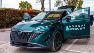 Fotografia em ângulo frontal de um SUV verde-escuro da marca Nissan estacionado em piso pavimentado. O veículo está com a porta do motorista e a tampa do porta-malas abertas, exibindo a inscrição lateral "QASHQAI e-POWER" em letras brancas. A grade dianteira ostenta o logotipo da montadora ao centro e faróis afilados em LED. Ao fundo, veem-se canteiros arborizados sob claridade diurna