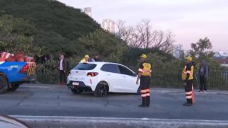 Fotografia ao amanhecer em cima de um viaduto mostrando um carro de passeio hatch branco atravessado na pista, com o pneu traseiro danificado. Dois agentes da CET (Companhia de Engenharia de Tráfego), vestindo bonés amarelos e coletes refletivos escuros com a marca da CET em amarelo, estão de pé ao lado do veículo orientando a ocorrência