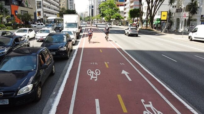 Fotografia em perspectiva frontal e iluminação diurna mostrando a ciclovia de canteiro central pintada em tom vermelho ao longo da Avenida Paulista. Em primeiro plano, a pista exibe pictogramas brancos de bicicleta e setas indicativas de sentido duplo. Ao fundo, dois ciclistas pedalam no trecho central. À esquerda, veículos de passeio e utilitários aguardam em fila no tráfego; à direita, a via rápida aparece desimpedida ao lado de um totem digital marcando a temperatura de 29°C, árvores e edifícios comerciais