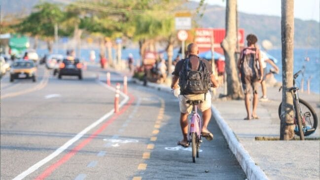Fotografia diurna em plano médio focando um ciclista visto de costas com mochila pilotando uma bicicleta roxa em uma ciclovia delimitada no asfalto. A pista exclusiva para bicicletas é separada da faixa de carros à esquerda por uma linha vermelha e balizadores verticais laranjas. À direita, pedestres caminham sobre o calçadão da orla ao lado do mar e de troncos de árvores sob luz solar fraca do fim de tarde