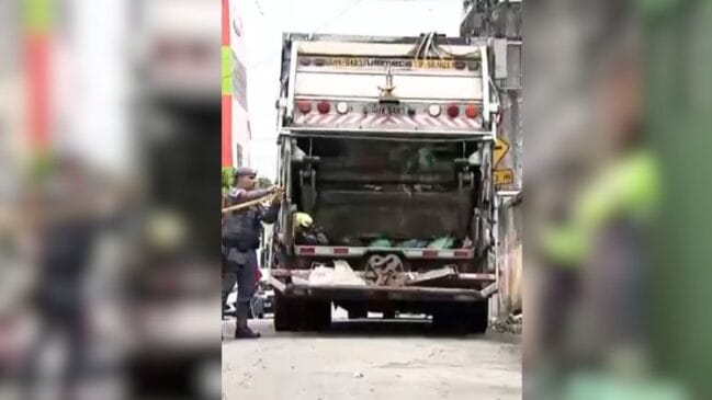 Fotografia diurna em plano médio focando a parte traseira de um caminhão de coleta de lixo cinza e branco estacionado em uma via pública. À esquerda do veículo, um policial militar de fardamento escuro passa uma fita amarela de isolamento policial para isolar o acesso ao compartimento de carga. Dentro da caçamba traseira do caminhão, observam-se sacos de lixo e resíduos descartados. O fundo exibe a parede de um imóvel e construções urbanas sob iluminação natural