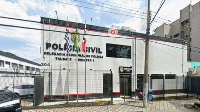 Fotografia de rua da fachada da Delegacia Seccional de Polícia de Taubaté (Deinter 1), da Polícia Civil do Estado de São Paulo. O prédio de fachada branca e detalhes em vermelho e preto tem o número 304 e o letreiro principal "POLÍCIA CIVIL" e "DELEGACIA SECCIONAL DE POLÍCIA TAUBATÉ - DEINTER 1". À frente, há mastros com as bandeiras do Brasil, São Paulo e Taubaté. Um pino de localização do Google Maps está no topo. À direita, placa indicando "Plantão 24h"