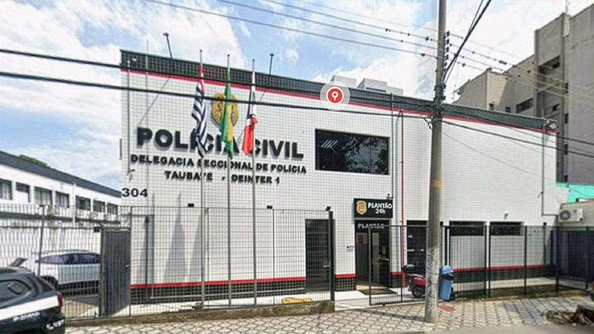 Fotografia de rua da fachada da Delegacia Seccional de Polícia de Taubaté (Deinter 1), da Polícia Civil do Estado de São Paulo. O prédio de fachada branca e detalhes em vermelho e preto tem o número 304 e o letreiro principal "POLÍCIA CIVIL" e "DELEGACIA SECCIONAL DE POLÍCIA TAUBATÉ - DEINTER 1". À frente, há mastros com as bandeiras do Brasil, São Paulo e Taubaté. Um pino de localização do Google Maps está no topo. À direita, placa indicando "Plantão 24h"