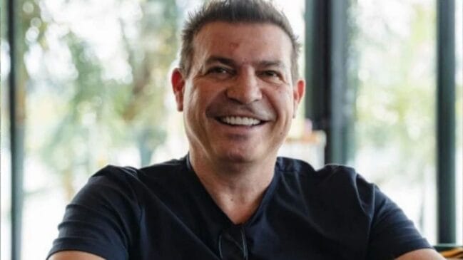 Fotografia diurna em plano médio de um homem de meia-idade sorrindo abertamente para a câmera. Ele possui cabelos curtos de cor castanho-escura com estilo arrepiado no topo, leve barba aparada e marcas de expressão ao redor dos olhos. Veste uma camiseta preta de gola careca e traz um par de óculos de armação escura enganchado no decote da blusa. O fundo da imagem apresenta uma estrutura de janelas de vidro com luz natural e vegetação verde desfocada ao fundo