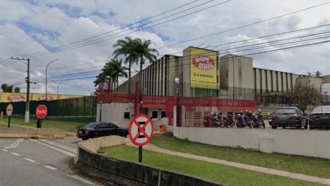 Fotografia no nível da rua mostrando a guarita de acesso e a fachada da fábrica da Bombril em São Bernardo do Campo. Em primeiro plano, vê-se a alça de acesso asfaltada com mureta de concreto e uma placa circular de trânsito indicando "Proibido Parar e Estacionar", além de uma placa de "PARE" à esquerda. Ao centro, a portaria possui portões vermelhos e guarita cinza. Ao fundo, destaca-se o edifício industrial com um grande painel amarelo exibindo o logotipo oval vermelho da Bombril e o slogan "É o máximo", ao lado de palmeiras altas e fiação elétrica