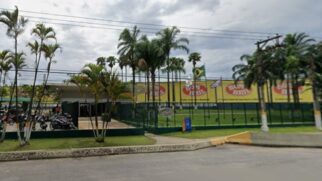 Vista frontal da entrada da fábrica da Bombril em São Bernardo do Campo. Em primeiro plano, veem-se a calçada com meio-fio, um canteiro com gramado verde, pequenas palmeiras e a guarita de acesso. Ao fundo, destaca-se o grande galpão industrial com fachada amarela estampada com múltiplos logotipos ovais vermelhos da marca "Bombril".