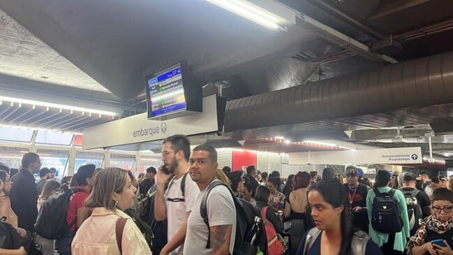 Fotografia em plano médio sob iluminação artificial no saguão de uma estação do Metrô de São Paulo repleta de passageiros com mochilas durante o horário de pico. No centro superior, suspenso no teto ao lado das placas de sinalização "embarque" e "Autoatendimento Poupatempo", um painel digital exibe o horário "07:40" e o status do sistema indicando "Velocidade Reduzida" para a Linha 1-Azul e a Linha 3-Vermelha, enquanto a Linha 2-Verde consta como "Operação Normal"