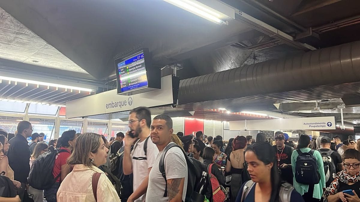Fotografia em plano médio sob iluminação artificial no saguão de uma estação do Metrô de São Paulo repleta de passageiros com mochilas durante o horário de pico. No centro superior, suspenso no teto ao lado das placas de sinalização "embarque" e "Autoatendimento Poupatempo", um painel digital exibe o horário "07:40" e o status do sistema indicando "Velocidade Reduzida" para a Linha 1-Azul e a Linha 3-Vermelha, enquanto a Linha 2-Verde consta como "Operação Normal"