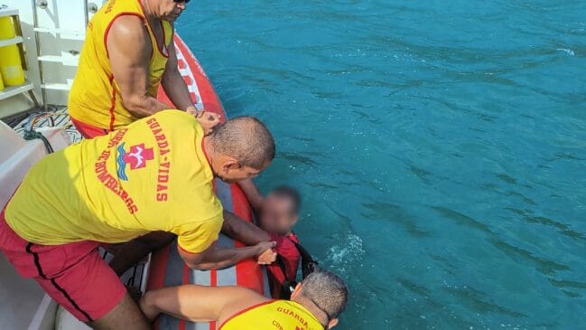 Fotografia em ângulo superior e iluminação diurna mostrando três guarda-vidas do Agrupamento de Bombeiros Marítimos (GBMar), vestindo regatas amarelas com a inscrição "Guarda-Vidas / Corpo de Bombeiros" e calções vermelhos, realizando o resgate de um homem na água. Os bombeiros estão posicionados na borda de um bote inflável e puxam o homem, que veste camisa vermelha e está com o rosto desfocado por sobreposição digital, para fora do mar de águas esverdeadas
