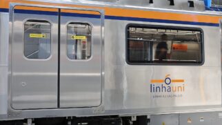 Foto de perfil da lateral de um trem de metrô de aço inox com listras superior amarela e azul. No corpo de metal, sobressai o logotipo "linhauni - LINHA 6 | SÃO PAULO". À esquerda, há uma porta dupla de acesso com visores de vidro e adesivos amarelos de alerta. Pela janela da composição, observa-se a plataforma iluminada da estação.