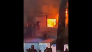 Fotografia noturna em baixa resolução mostrando um incêndio de grandes proporções em um imóvel no Guarujá. No andar superior, chamas intensas nas cores laranja e amarelo são visíveis através de uma grande janela de caixilharia escurecida. No pavimento inferior, veem-se as silhuetas de pessoas e socorristas observando o fogo, acompanhadas por reflexos de luzes de emergência azuis e vermelhas. Um poste escuro corta verticalmente a imagem na parte direita