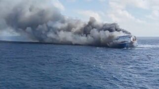 Fotografia em plano aberto no oceano mostrando um navio de passageiros/ferry atingido por um incêndio em alto-mar. Uma densa e espessa cortina de fumaça cinza-escura e preta sai do corpo da embarcação e se alastra para a esquerda sobre a superfície do mar azul sob céu claro.