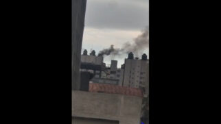 Fotografia na vertical tirada à distância mostrando uma coluna de fumaça escura e densa subindo do topo de um edifício comercial. A fumaça emerge de trás das estruturas de caixa-d'água e do topo do prédio, dispersando-se sob um céu cinzento e totalmente coberto por nuvens. Em primeiro plano, aparecem partes do topo e telhados de construções vizinhas.