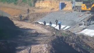 Fotografia em ângulo elevado de uma área de obras e terraplenagem no perímetro do Aeroporto Internacional de Guarulhos (GRU Airport). O terreno exibe solo exposto, cortes na terra e uma rampa com camada de brita e telas de contenção. Na parte superior à direita, há maquinário pesado amarelo (caminhão/escavadeira) e um banheiro químico azul. Ao longo do barranco e do caminho de terra, veem-se várias pessoas com roupas escuras transitando pelo local