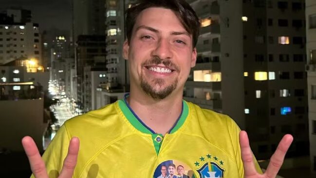 Descrição curta “para cego ver”: Homem sorridente com camisa amarela da seleção brasileira, fazendo sinal de paz com as mãos, à noite, em varanda com prédios iluminados ao fundo.