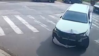 Carro branco com a frente muito danificada, parado próximo a uma faixa de pedestres em rua asfaltada, com outros veículos e árvores ao fundo.