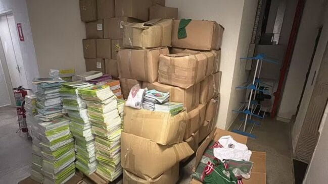 Fotografia em iluminação interna mostrando pilhas de cadernos e livros didáticos empilhados diretamente no chão, ao lado de dezenas de caixas de papelão fechadas e abertas contendo uniformes escolares nas cores branca e verde. O material está acumulado no corredor de um depósito ou prédio público, próximo a extintores de incêndio e uma arara de roupas vazia