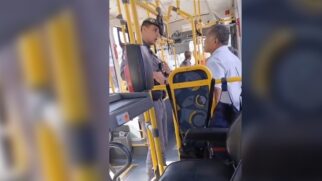 Fotografia na vertical capturada do interior de um ônibus coletivo urbano mostrando um policial militar fardado e de boina escura em pé no corredor. Ele conversa de frente e a curta distância com um homem de cabelos grisalhos que veste camisa social clara de mangas curtas