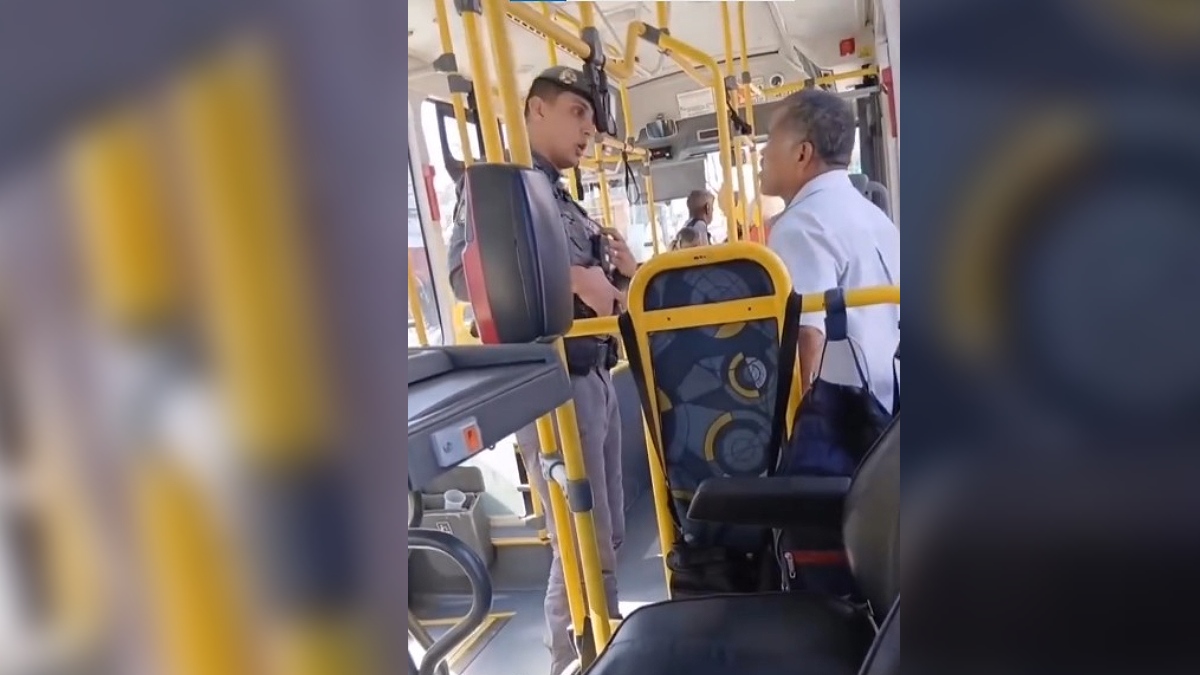 Fotografia na vertical capturada do interior de um ônibus coletivo urbano mostrando um policial militar fardado e de boina escura em pé no corredor. Ele conversa de frente e a curta distância com um homem de cabelos grisalhos que veste camisa social clara de mangas curtas